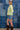 A woman wearing a lime green ribbed long-sleeve crewneck sweater with 'TEXAS' printed in white block letters across the chest, paired with denim shorts and a camouflage baseball cap, standing against a blue denim patchwork backdrop-Fleastyle