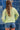 A woman wearing a lime green ribbed long-sleeve crewneck sweater with 'TEXAS' printed in white block letters across the chest, paired with denim shorts and a camouflage baseball cap, standing against a blue denim patchwork backdrop-Fleastyle
