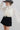 A woman wearing a cream long-sleeve dramatic blouse with black polka dots and a high gathered neckline and adjustable neck tie, paired with a black mini skort and a gray cowboy hat, against a light gray studio background--Flowy_Polka_Dot_Tie_Blouse_-_Ivory-FleaStyle  