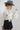 A woman wearing a cream long-sleeve dramatic blouse with black polka dots and a high gathered neckline and adjustable neck tie, paired with a black mini skort and a gray cowboy hat, and ivory cowboy boots against a light gray studio background--Flowy_Polka_Dot_Tie_Blouse_-_Ivory-FleaStyle  