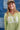 A woman wearing a lime green ribbed long-sleeve crewneck sweater with 'TEXAS' printed in white block letters across the chest, paired with denim shorts and a camouflage baseball cap, standing against a blue denim patchwork backdrop-Fleastyle