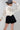A woman wearing a cream long-sleeve dramatic blouse with black polka dots and a high gathered neckline and adjustable neck tie, paired with a black mini skort  with a flattering high-rise fit with a flowy silhouette and a gray cowboy hat, against a light gray studio background--Suede Pleated Skort - Black