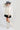 A woman wearing a cream long-sleeve dramatic blouse with black polka dots and a high gathered neckline and adjustable neck tie, paired with a black mini skort and a gray cowboy hat, and ivory cowboy boots against a light gray studio background--Flowy_Polka_Dot_Tie_Blouse_-_Ivory-FleaStyle  
