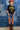 A woman wearing a black graphic t-shirt with yellow 'FORT WORTH TEXAS' lettering, paired with denim shorts, a rust cowboy hat, and brown cowboy boots, standing in front of a blue denim patchwork backdrop-Fleastyle.