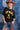 A woman wearing a black graphic t-shirt with yellow 'FORT WORTH TEXAS' lettering, paired with denim shorts, a rust cowboy hat, and brown cowboy boots, standing in front of a blue denim patchwork backdrop-Fleastyle.