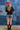 A woman wearing a black graphic t-shirt with yellow 'FORT WORTH TEXAS' lettering, paired with denim shorts, a rust cowboy hat, and brown cowboy boots, standing in front of a blue denim patchwork backdrop-Fleastyle.