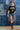 A woman wearing a black graphic t-shirt with yellow 'FORT WORTH TEXAS' lettering, paired with denim shorts, a rust cowboy hat, and brown cowboy boots, standing in front of a blue denim patchwork backdrop-Fleastyle.