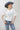 A woman wearing a white short-sleeve crop top with a lace sheer underlay in a delicate floral pattern, with fitted silhouette and thumb holes, blue jeans, and a dusty blue cowboy hat -Lace_Mock_Neck_Sheer_Underlay_-_Beige-FleaStyle