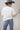 A woman wearing a white short-sleeve crop top with a lace sheer underlay in a delicate floral pattern, with fitted silhouette and thumb holes, blue jeans, and a dusty blue cowboy hat -Lace_Mock_Neck_Sheer_Underlay_-_Beige-FleaStyle