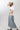 A woman wearing a white graphic t-shirt with brown western-style text reading 'Y'ALL NEED DOLLY', styled with a beige cowboy hat, sheer patterned shirt, and light blue jeans against a plain light gray background-Y_all_Need_Dolly_Graphic_T-Shirt-FleaStyle
