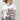 A woman wearing a white graphic t-shirt with brown western-style text reading 'Y'ALL NEED DOLLY', styled with a beige cowboy hat, sheer patterned shirt, and light blue jeans against a plain light gray background-Y_all_Need_Dolly_Graphic_T-Shirt.