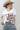 A woman wearing a white graphic t-shirt with brown western-style text reading 'Y'ALL NEED DOLLY', styled with a beige cowboy hat, sheer patterned shirt, and light blue jeans against a plain light gray background-Y_all_Need_Dolly_Graphic_T-Shirt.
