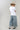 A woman wearing a white graphic t-shirt with brown western-style text reading 'Y'ALL NEED DOLLY', styled with a beige cowboy hat, sheer patterned shirt, and light blue jeans against a plain light gray background-Y_all_Need_Dolly_Graphic_T-Shirt-FleaStyle