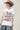 A woman wearing a white graphic t-shirt with brown western-style text reading 'Y'ALL NEED DOLLY', styled with a beige cowboy hat, sheer patterned shirt, and light blue jeans against a plain light gray background-Y_all_Need_Dolly_Graphic_T-Shirt-FleaStyle