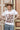 A woman wearing a white graphic t-shirt with brown western-style text reading 'Y'ALL NEED DOLLY', styled with a beige cowboy hat, sheer patterned shirt, and light blue jeans against a plain light gray background-Y_all_Need_Dolly_Graphic_T-Shirt-FleaStyle