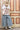A woman wearing a white graphic t-shirt with brown western-style text reading 'Y'ALL NEED DOLLY', styled with a beige cowboy hat, sheer patterned shirt, and light blue jeans against a plain light gray background-Y_all_Need_Dolly_Graphic_T-Shirt-FleaStyle