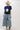 A woman wearing a classic mock neckline knit sweater, easy short sleeves and a bold ivory star, a beige cowboy hat, light-wash denim pants, and brown cowboy boots with blue star embroidery, sitting on a white studio floor--Star_Short-Sleeve_Knit_Sweater_-_Navy-FleaStyle