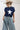 A woman wearing a classic mock neckline knit sweater, easy short sleeves and a bold ivory star, a beige cowboy hat, light-wash denim pants, and brown cowboy boots with blue star embroidery, sitting on a white studio floor--Star_Short-Sleeve_Knit_Sweater_-_Navy-FleaStyle