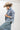 A woman wearing a relaxed, light-wash distressed denim jacket with raw frayed hem covered in shimmering rhinestones for a playful take on a forever classic ,  paired with wide-leg denim pants, and a beige cowboy hat  posed against a plain light gray studio background--Raw_Hem_Bling_Denim_Shacket-FleaStyle