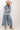A woman wearing a relaxed, light-wash distressed denim jacket with raw frayed hem covered in shimmering rhinestones for a playful take on a forever classic ,  paired with wide-leg denim pants, and a beige cowboy hat  posed against a plain light gray studio background--Raw_Hem_Bling_Denim_Shacket-FleaStyle