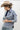 A woman wearing a relaxed, light-wash distressed denim jacket with raw frayed hem covered in shimmering rhinestones for a playful take on a forever classic ,  paired with wide-leg denim pants, and a beige cowboy hat  posed against a plain light gray studio background--Raw_Hem_Bling_Denim_Shacket-FleaStyle