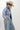 A woman wearing a relaxed, light-wash distressed denim jacket with raw frayed hem covered in shimmering rhinestones for a playful take on a forever classic ,  paired with wide-leg denim pants, and a beige cowboy hat  posed against a plain light gray studio background--Raw_Hem_Bling_Denim_Shacket-FleaStyle