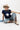 A woman wearing a classic mock neckline knit sweater, easy short sleeves and a bold ivory star, a beige cowboy hat, light-wash denim pants, and brown cowboy boots with blue star embroidery, sitting on a white studio floor--Star_Short-Sleeve_Knit_Sweater_-_Navy-FleaStyle