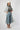 A woman modeling a bohemian-style outfit featuring a cream crochet kimono with blue floral patterns, a brown graphic t-shirt, light-wash denim culottes, a white cowboy hat, and brown lace-up heeled sandals against a light gray studio background.