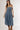A woman wearing a strapless tiered denim maxi dress in a medium blue wash, paired with a brown baseball cap that reads 'Laguna BEACH' and layered gold necklaces-Fleastyle