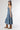 A woman wearing a strapless tiered denim maxi dress in a medium blue wash, paired with a brown baseball cap that reads 'Laguna BEACH' and layered gold necklaces-Fleastyle