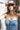 A woman wearing a strapless tiered denim maxi dress in a medium blue wash, paired with a brown baseball cap that reads 'Laguna BEACH' and layered gold necklaces-Fleastyle