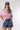 A woman wearing soft pink striped pattern, relaxed boxy silhouette and easy half sleevesshirt, paired with blue denim shorts, brown cowboy boots, and a navy blue baseball cap with a -FleaStyle