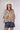 A woman modeling a bohemian-style long-sleeve button-up blouse with a multicolor paisley and floral print on a cream base, paired with blue bandana-print shorts and a white cowboy hat, against a light gray studio background.