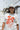 A woman stands on a beach wearing a ivory graphic tee featuring a relaxed fit with bold California orange poppies, denim shorts, and a beige baseball cap, with ocean waves and rocky shoreline in the background--California_Poppy_Oversized_Graphic_T-Shirt_-_Ivory_Orange-Fleastyle.