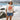 A woman stands on a beach wearing a ivory graphic tee featuring a relaxed fit with bold California orange poppies, denim shorts, and a beige baseball cap, with ocean waves and rocky shoreline in the background--California_Poppy_Oversized_Graphic_T-Shirt_-_Ivory_Orange-Fleastyle.