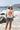A woman stands on a beach wearing a ivory graphic tee featuring a relaxed fit with bold California orange poppies, denim shorts, and a beige baseball cap, with ocean waves and rocky shoreline in the background--California_Poppy_Oversized_Graphic_T-Shirt_-_Ivory_Orange-Fleastyle.