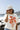 A woman stands on a beach wearing a ivory graphic tee featuring a relaxed fit with bold California orange poppies, denim shorts, and a beige baseball cap, with ocean waves and rocky shoreline in the background--California_Poppy_Oversized_Graphic_T-Shirt_-_Ivory_Orange-Fleastyle.