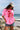 A woman standing on a rocky beach wearing a bright pink t-shirt with a red poppy graphic, denim shorts, and a beige baseball cap with 'LOCAL' written on it-Local Textured Patch Trucker Hat - Neutral-FleaStyle