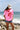 A woman standing on a rocky beach wearing a bright pink t-shirt with a red poppy graphic, denim shorts, and a beige baseball cap with 'LOCAL' written on it-Local Textured Patch Trucker Hat - Neutral