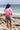 A woman stands on a rocky beach facing the ocean, wearing a bright pink t-shirt with a red floral poppy graphic and the word 'California' on the back, paired with denim shorts and a Roger-Cowboy-Straw-Hat-Black-FleaStyle