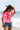 A woman standing on a beach wearing a bright pink t-shirt with a red poppy graphic, denim shorts, and a Roger-Cowboy-Straw-Hat-Black-FleaStyle