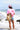 A woman stands on a beach facing the ocean, wearing a bright pink t-shirt with red lipstick marks and the word 'California' on the back, denim shorts, a pink trucker hat, and carrying a straw hat on her shoulder. California Poppy Oversized Graphic T-Shirt - Pink/Red Flea Style