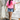 A woman walking down white outdoor stairs wearing a bright pink graphic t-shirt with red flowers, denim shorts, a black baseball cap with 'CALI' in white, and carrying a woven straw bag_ California Poppy Oversized Graphic T-Shirt - Pink/Red_Fleastyle