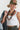 A woman wearing a white ribbed tank top and camouflage pants, an olive green trucker hat with a butterfly patch styled with A collected mix of olive green beads, dark stacked disc details, aged metal accents, and an antique gold-tone medallion pendant-Vintage_Tribal_Coin_Beaded_Necklace-FleaStyle 